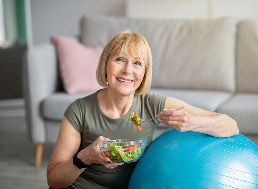 A woman smiles as she leans on an exercise ball and eats a salad.